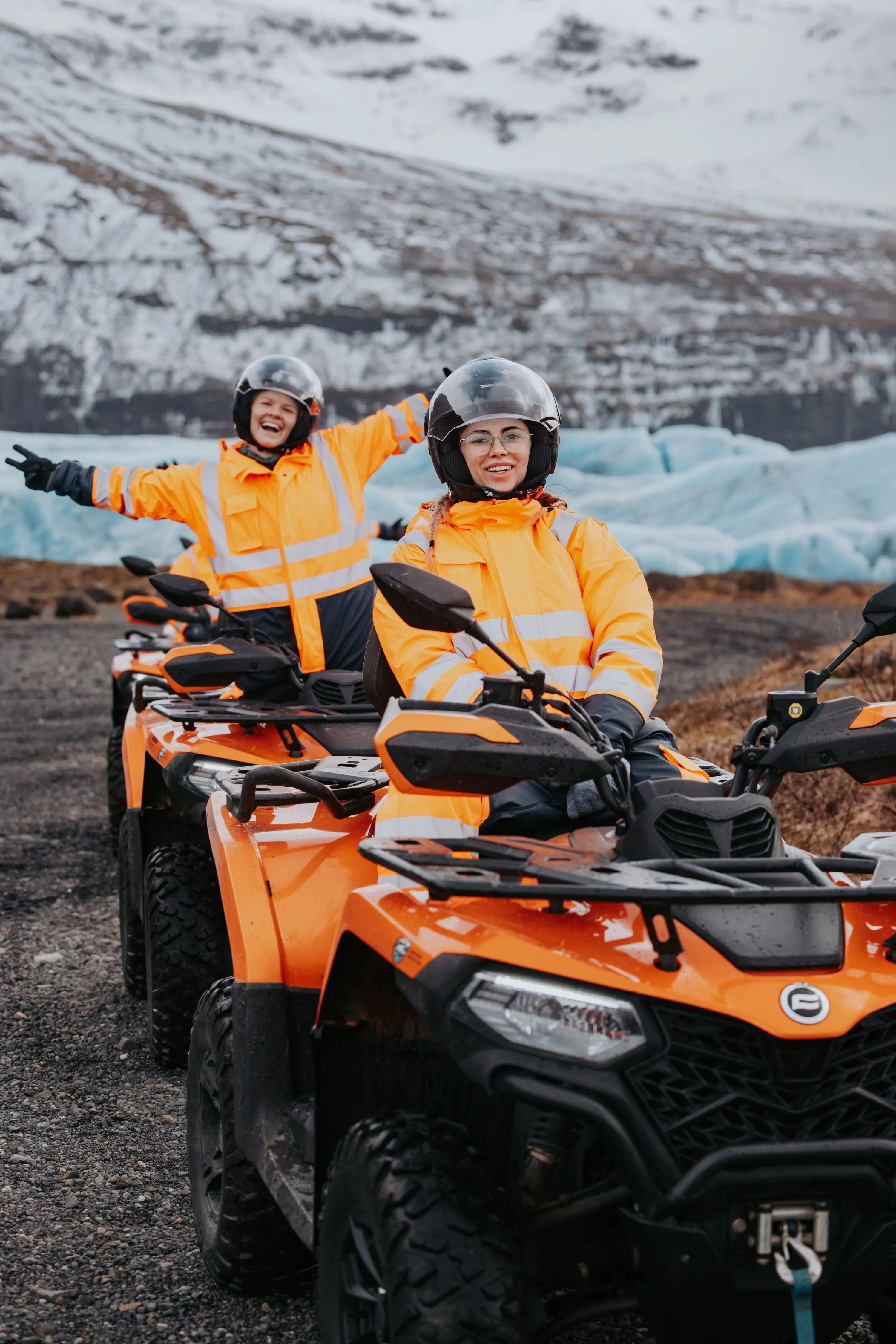 Two ATV riders in bright orange coveralls pose excitedly in front of a vibrant blue glacial outlet in winter.