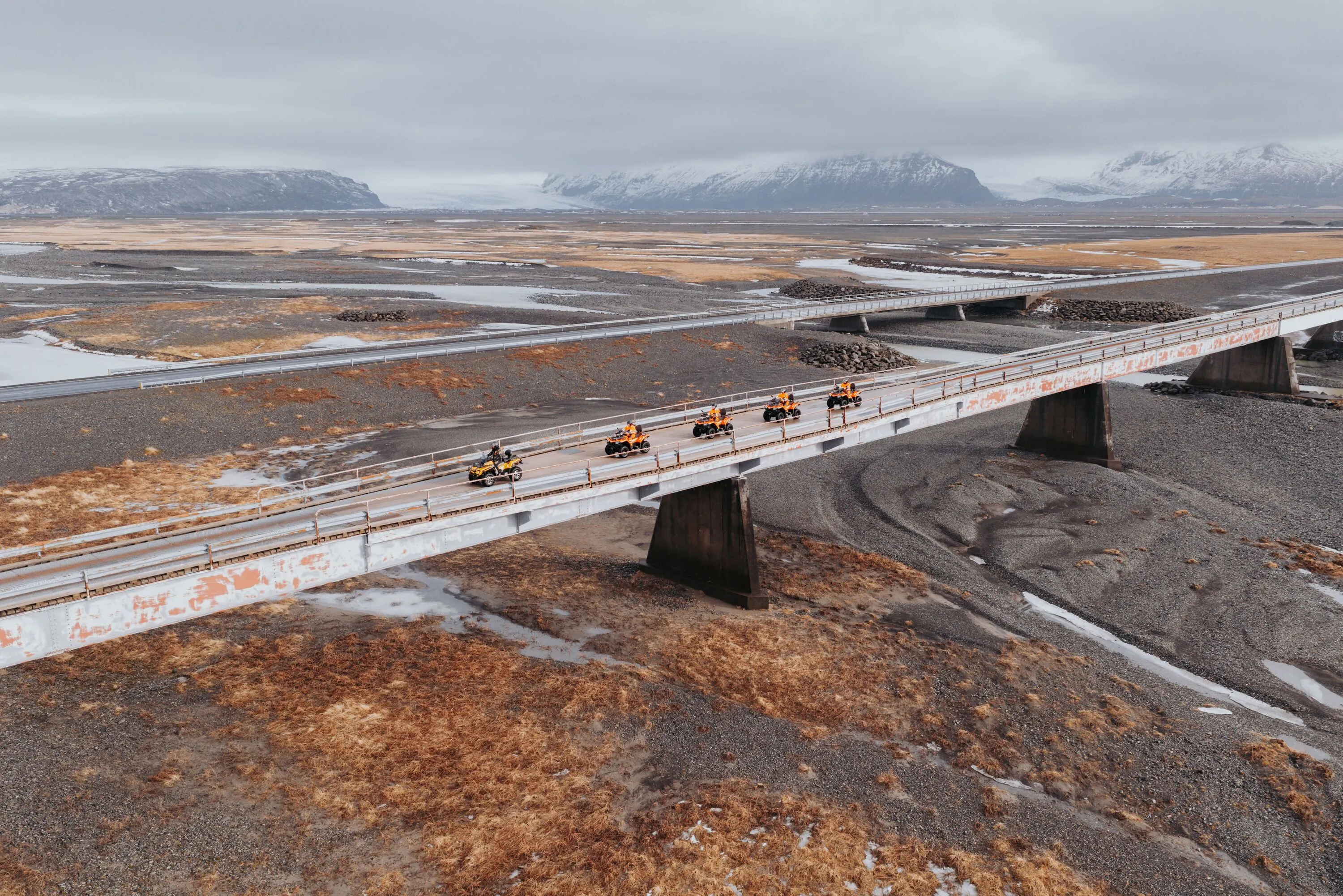 Riders on ATVs cross a long bridge over a dried up glacial river bed with glacier covered mountains in the background