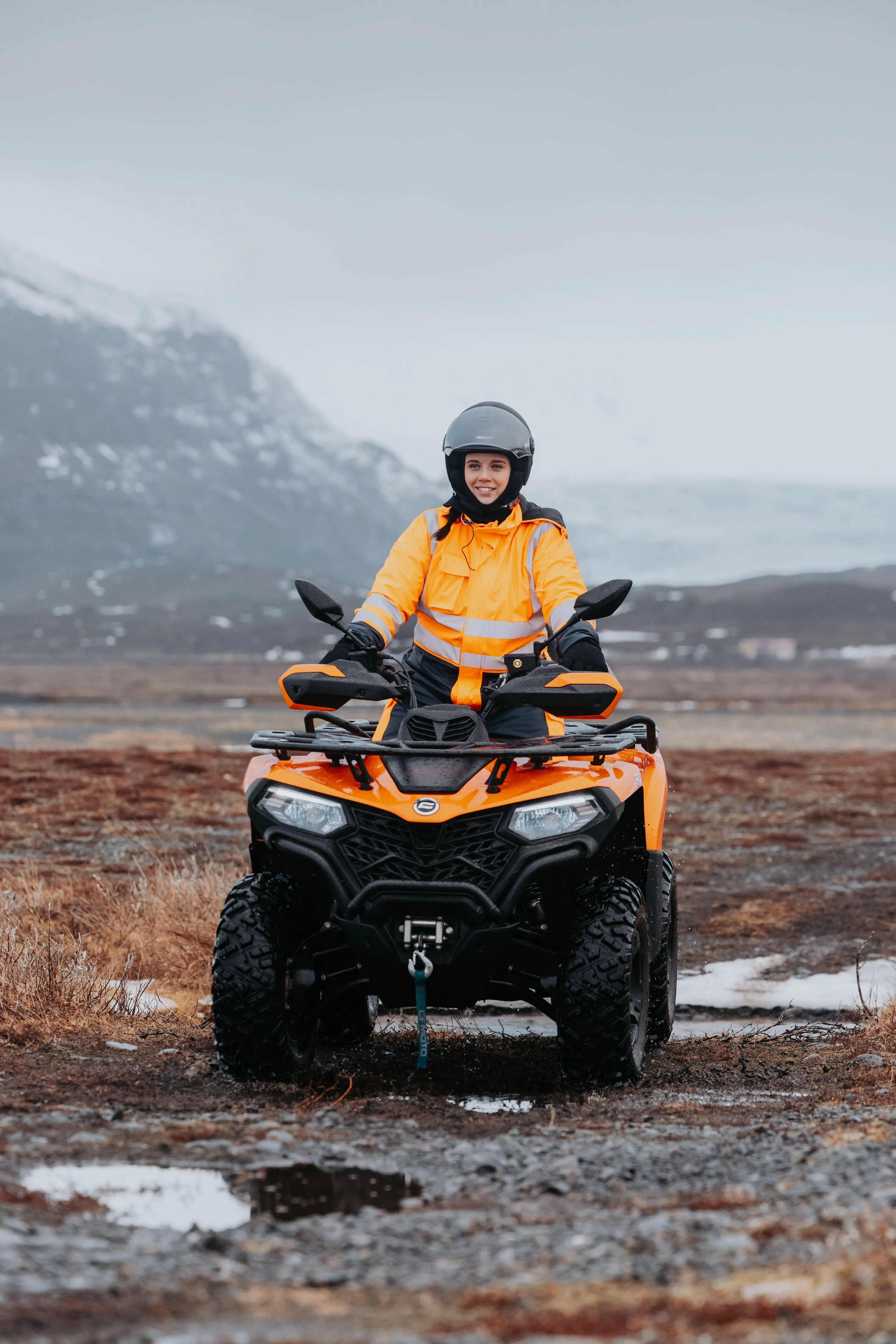 A woman on an ATV rides on a gravel track on a misty morning with mountains in the background.
