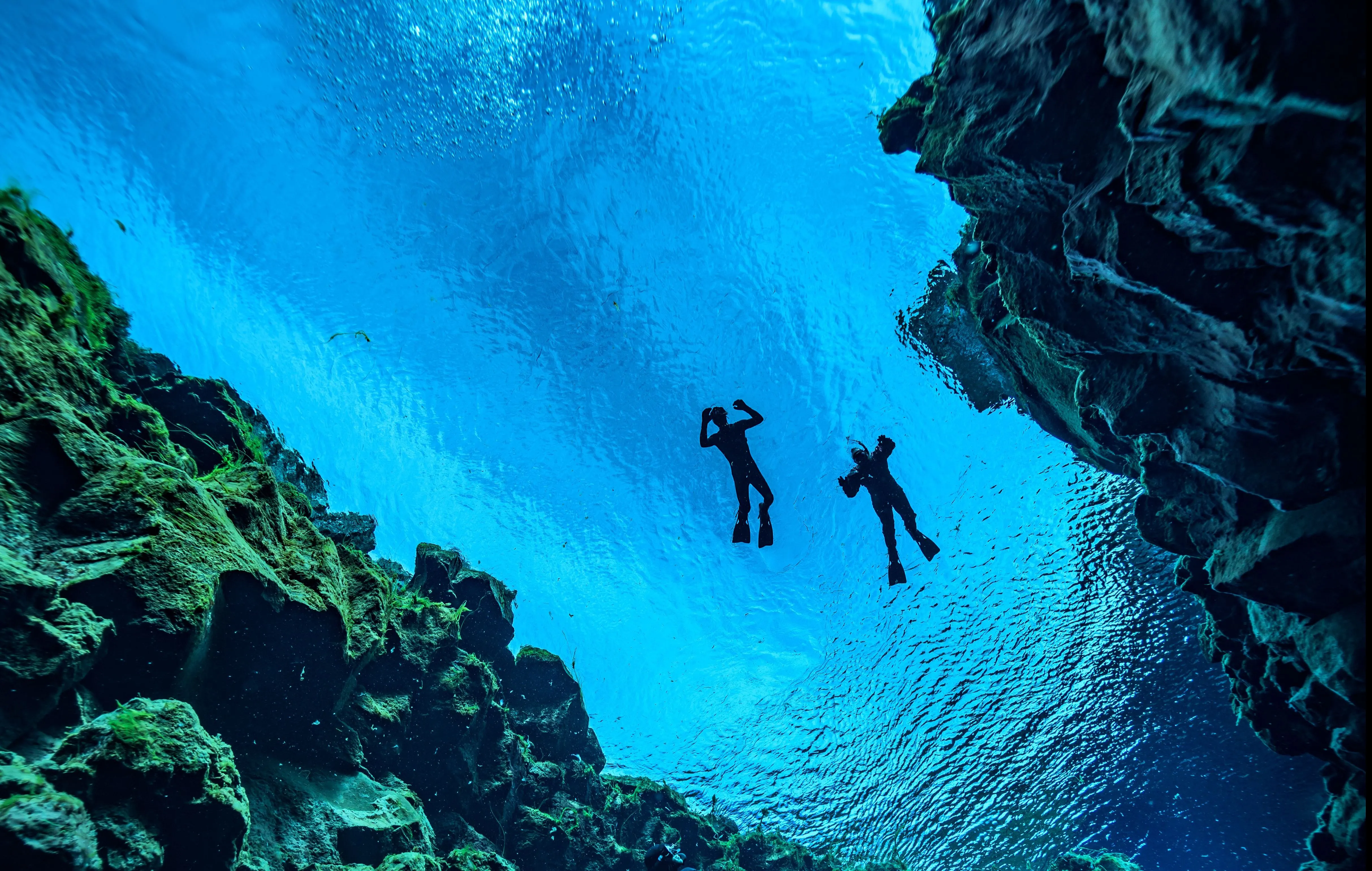 Sillouettes of two freedivers up on the surface of Silfra's vibrant blue waters between rocky fissure walls.