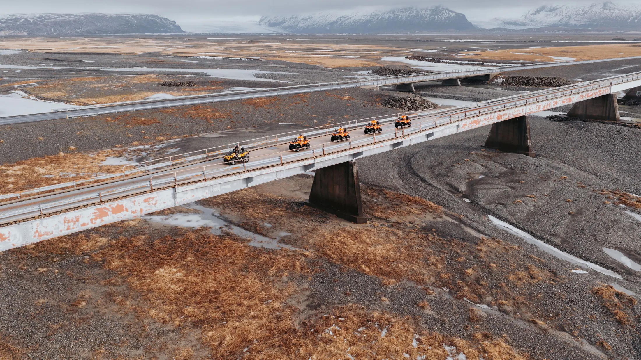Riders on ATVs cross a long bridge over a dried up glacial river bed with glacier covered mountains in the background
