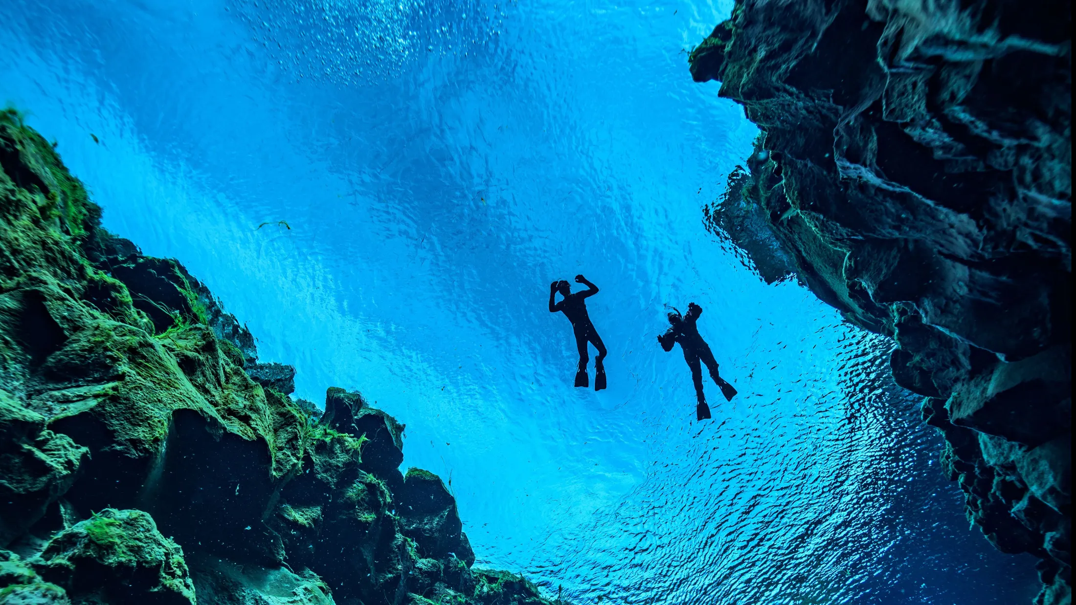 Sillouettes of two freedivers up on the surface of Silfra's vibrant blue waters between rocky fissure walls.