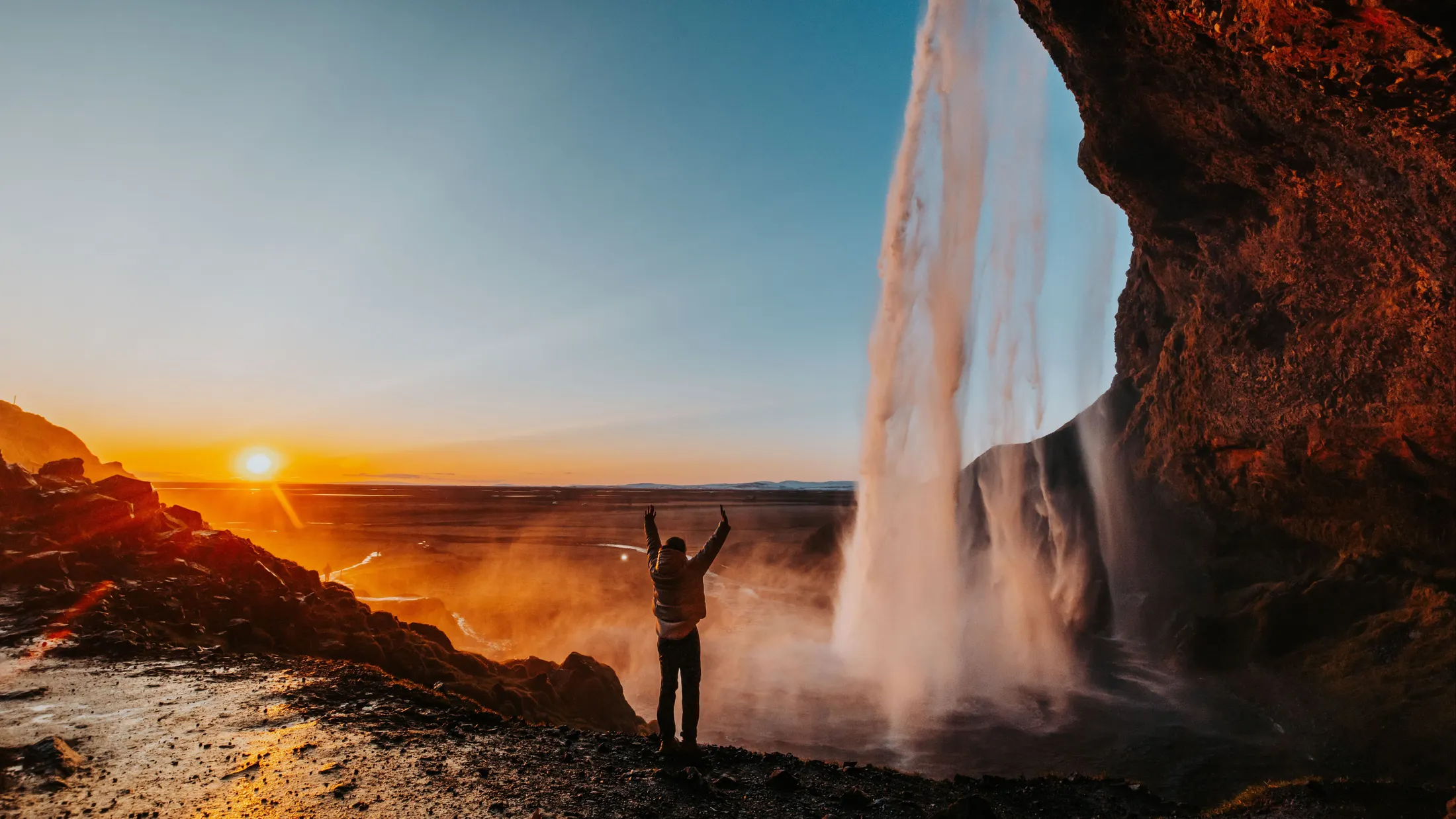 A sillouette of a person with their hands up stands next to a waterfall pouring over an overhanging cliff at sunset.