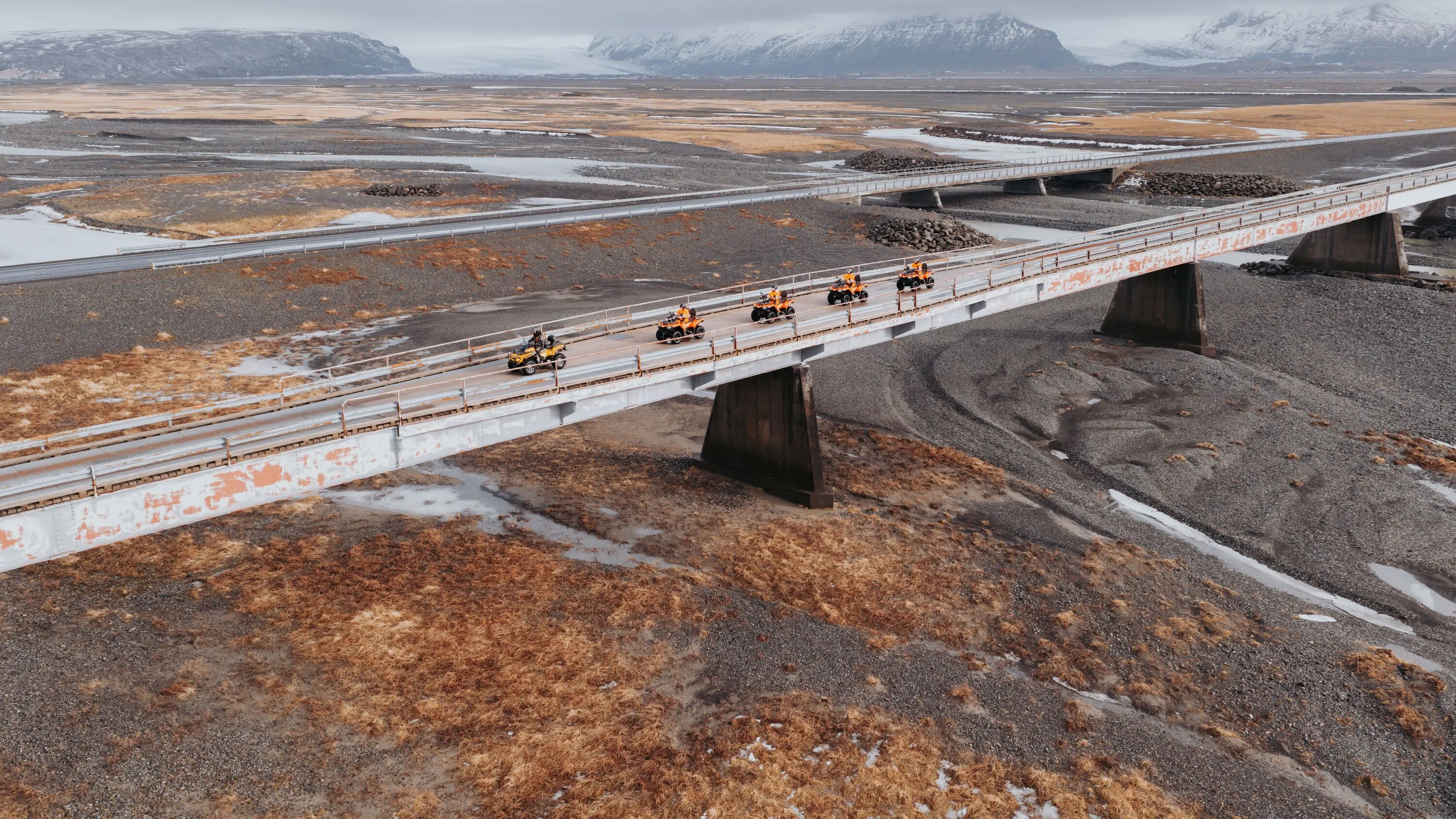 Riders on ATVs cross a long bridge over a dried up glacial river bed with glacier covered mountains in the background