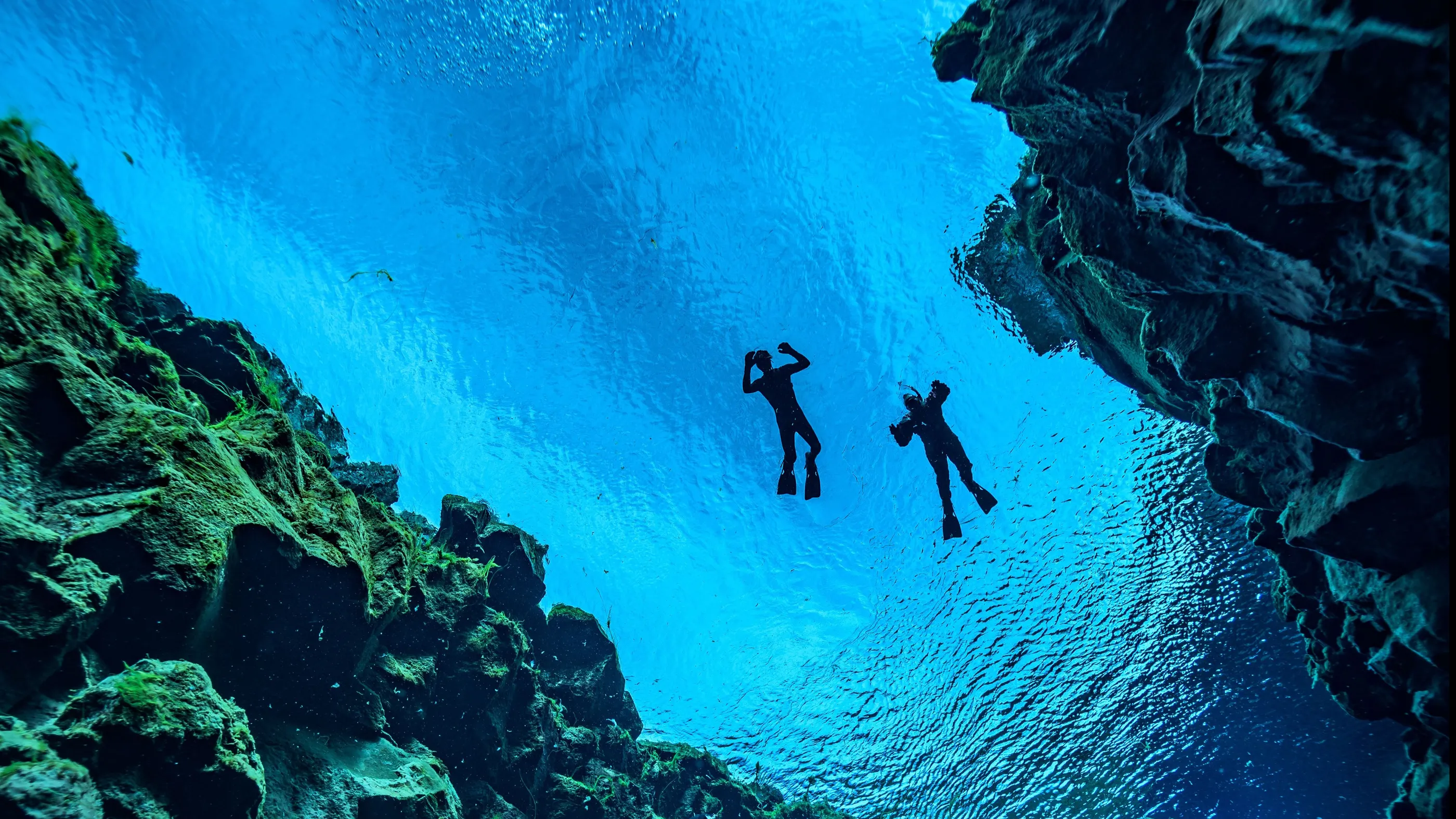 Sillouettes of two freedivers up on the surface of Silfra's vibrant blue waters between rocky fissure walls.