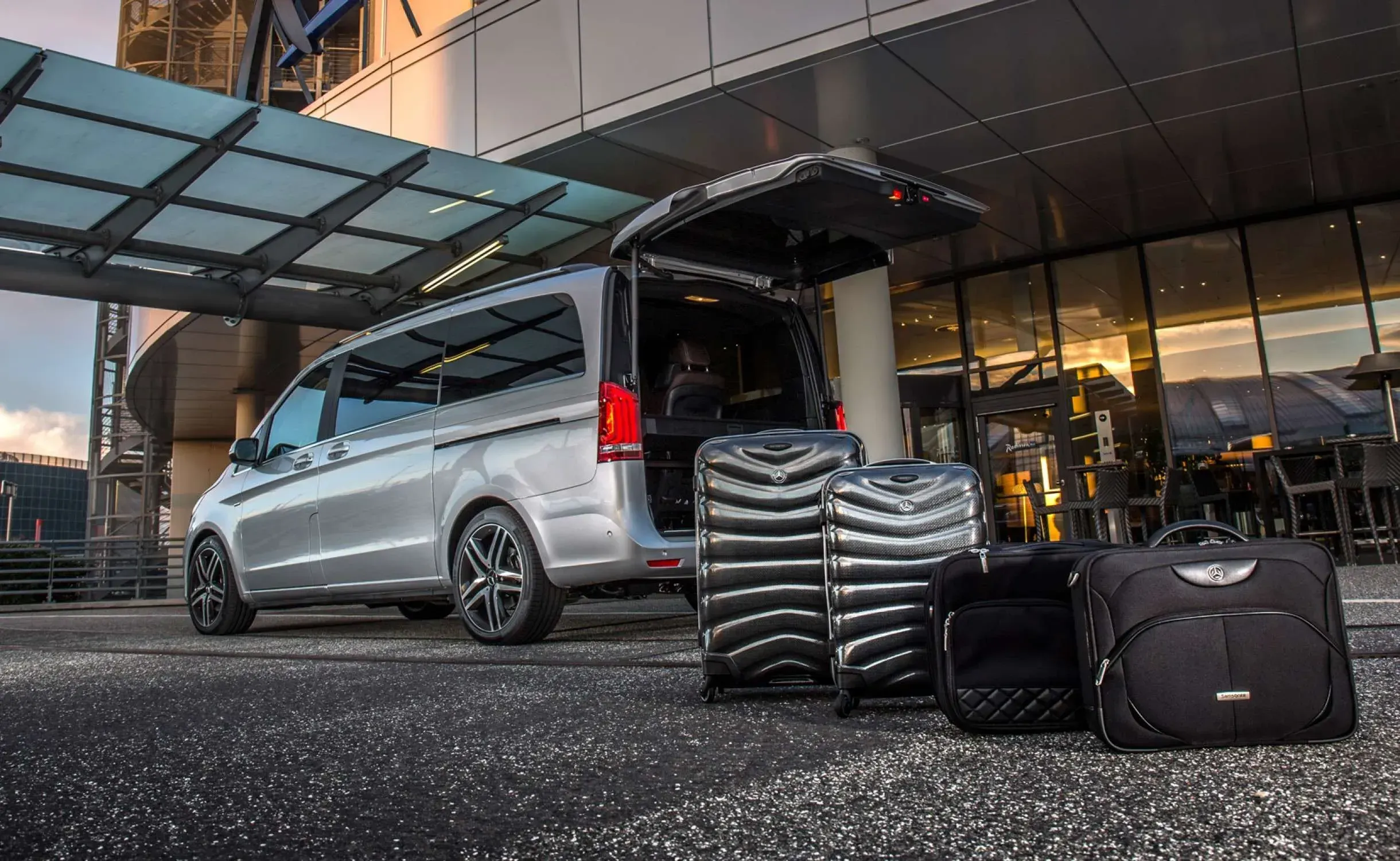 A silver van with an open trunk sits outside Keflavík airport and suitcases waiting to be loaded 