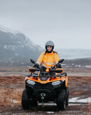 A woman on an ATV rides on a gravel track on a misty morning with mountains in the background.