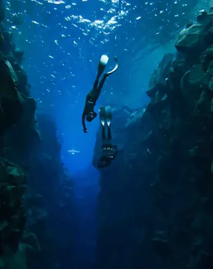 Two freedivers dive deeper into a fissure filled with clear, deep blue water.