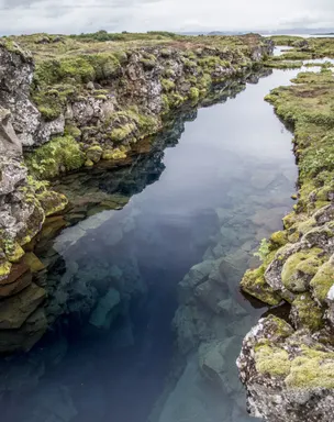 A long, narrow water- filled rift surrounded by moss covered rocks reflects sun on a partially cloudy day