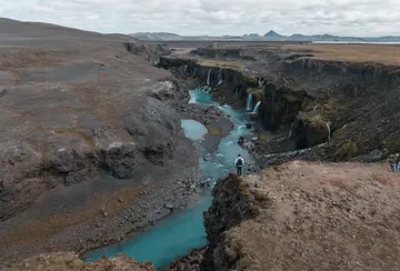 A person stands at a brown canyon edge overlooking the vibrant blue river flowing through it, with waterfalls pouring over the sides. 