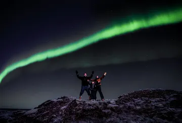 A family of three on a Northern Lights tour stand on a snowy hill with their arms up under a single vibrant green aurora line.