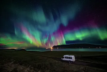 The 4x4 Super truck parks next to a lake, while very colorful northern lights explode across the sky.