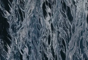 Hundreds of small, light blue glacial river streams flow over deep black volcanic sand in a unique weave-like pattern.