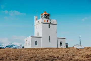The white rectangular lighthouse with an orange dome at Dyrhólaey viewpoint, with Mýrdalsjökull ice cap in the background on a clear day.