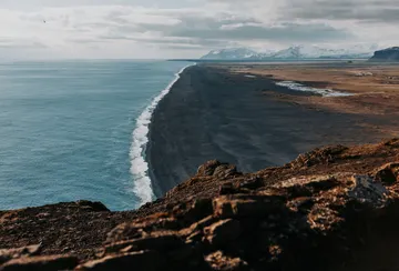 The pale blue ocean contrasts with Iceland's black volcanic coastline, with the white line of waves crashing along the shore and disappearing into the horizon.