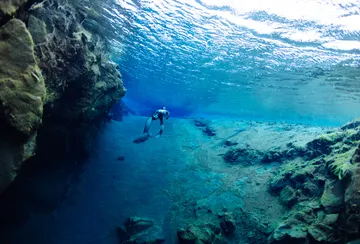 Sun sparkles on the surface of the water, as a free diver explores below along a fissure wall and sandy bottom.