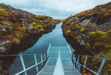 Metal stairs lead into the narrow Sifra fissure framed by moss- covered rocky walls on each side and the water is very still, reflecting the cloudy skies above.