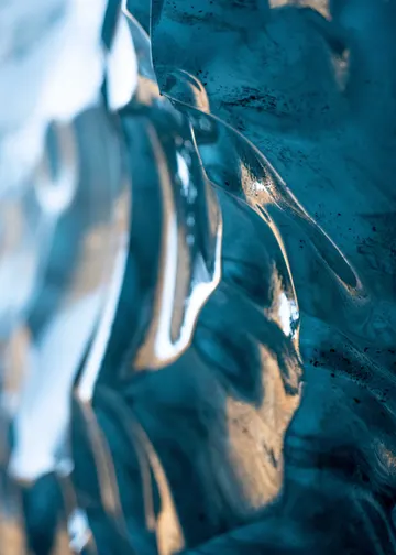 A vibrant blue glacial ice wall looks like ocean waves made of glass shimmering with warm, yellow sunlight.