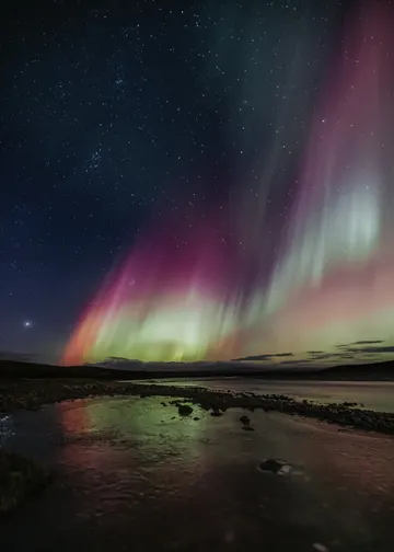 A faint glow of green and purple northern lights are reflected in a stream as they dance above in the dark sky.