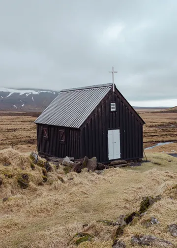 A small, old church constructed of dark wood sits on a light yellow meadow with lightly snow covered mountains in the background on an autumn day.