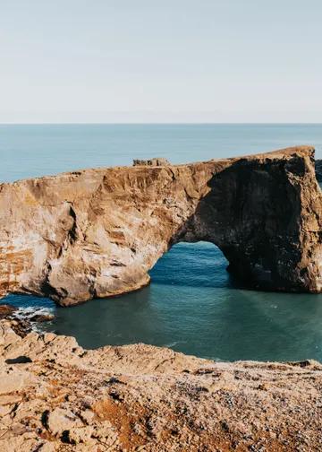 A promontory juts out into the ocean, ending with a massive rock arch on a sunny day.