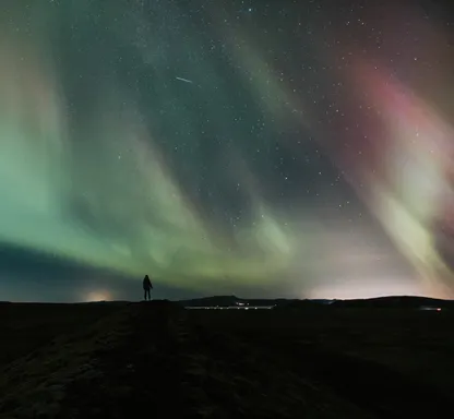 A person's silhouette appears very small under a sky completely covered with light green Northern lights.