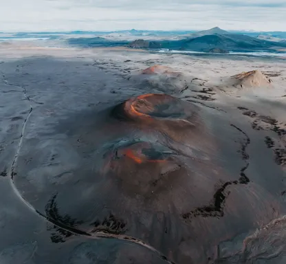 Three volcanoes with orange rims in the middle of ash- brown flat land, with mountains in the distant background.