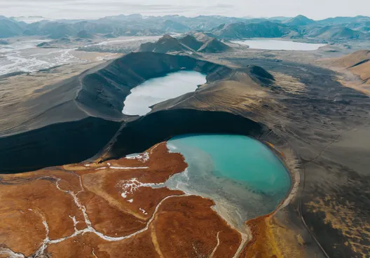 Vibrant rusty-orange terrain leads to a turquoise colored lake in a volcanic crater, in front of a second crater lake.