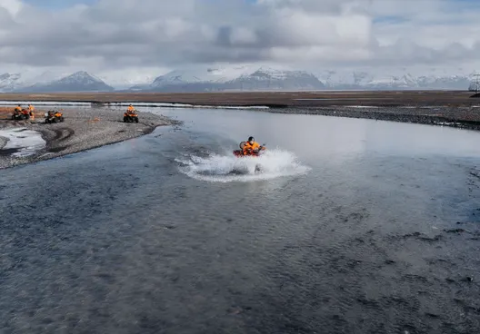 An ATV rider makes a big splash as they cross a wide, clear river, the sun gives it a light blue appearance.