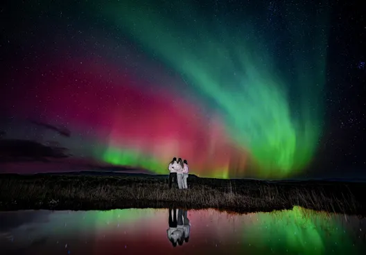Three people on a small group Northern Lights tour stand on the edge of a pond under a vibrant aurora.