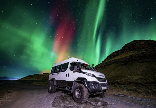 On tour, the 4x4 vehicle called the Super Truck is parked next to a small grassy mountain while the Northern lights look like a colorful explosion in the sky.