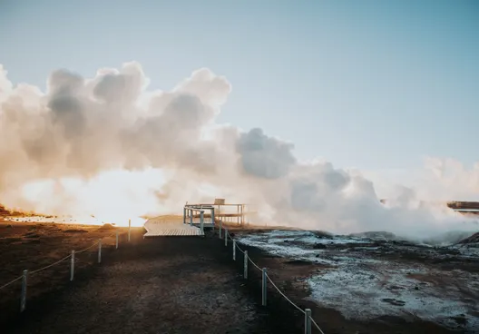 Geothermal steam billows, creating puffy white clouds that float over a walkway as the sun begins to set.