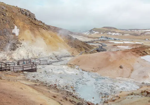 Tourists on a long, wooden walkway observe geothermal steam rising up from light- grey muddy puddles surrounded by tan, peach and rust-colored hilly terrain.