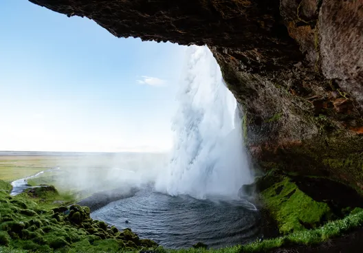 View from the back of Seljalandsfoss pouring into a small pool as it sprays mist, partially concealing the green field in the background.
