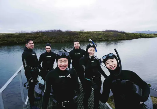 A group of happy tourists wearing wetsuits, dive masks and snorkels stand smiling in front of Silfra after the free diving tour.