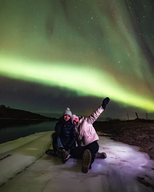 Two people sit on the icy ground below a sky lit up by a light green aurora.