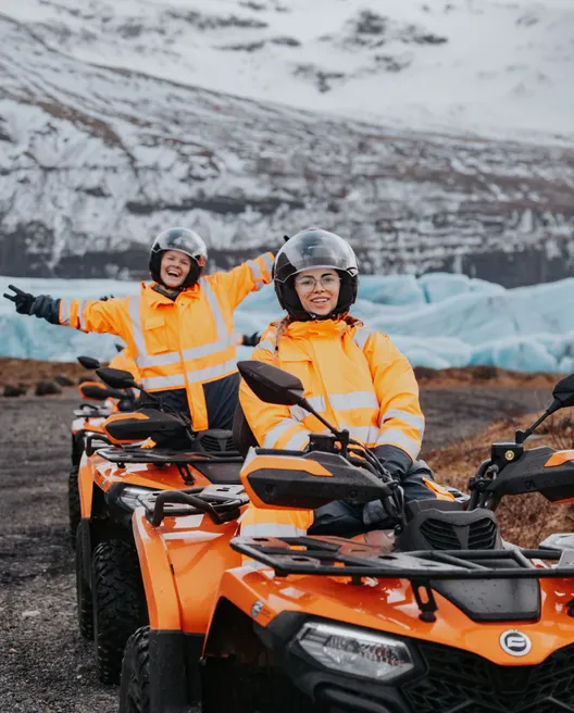 Two ATV riders in bright orange coveralls pose excitedly in front of a vibrant blue glacial outlet in winter.