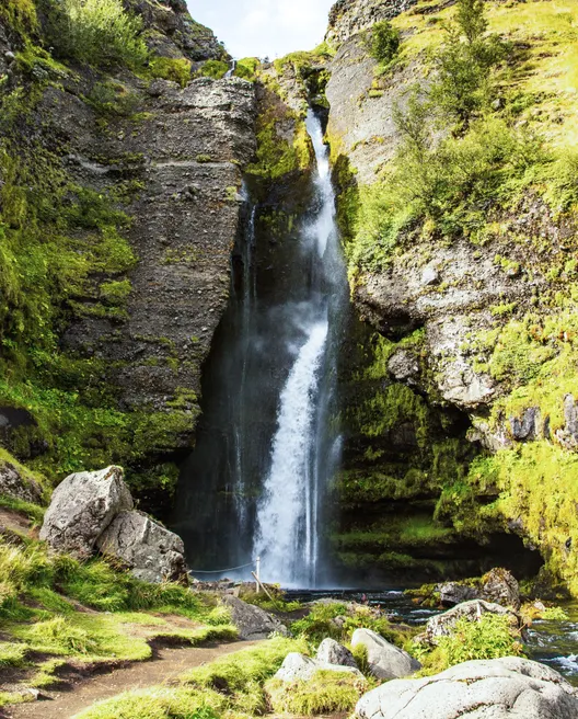 A partially concealed waterfall flows between layered rock formations covered in green moss on a sunny day.