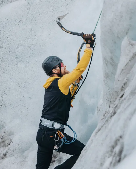 A client in a yellow jacket and black helmet holds his ice axe, about to take a swing at the base of a white glacial wall and begin the ice climb.