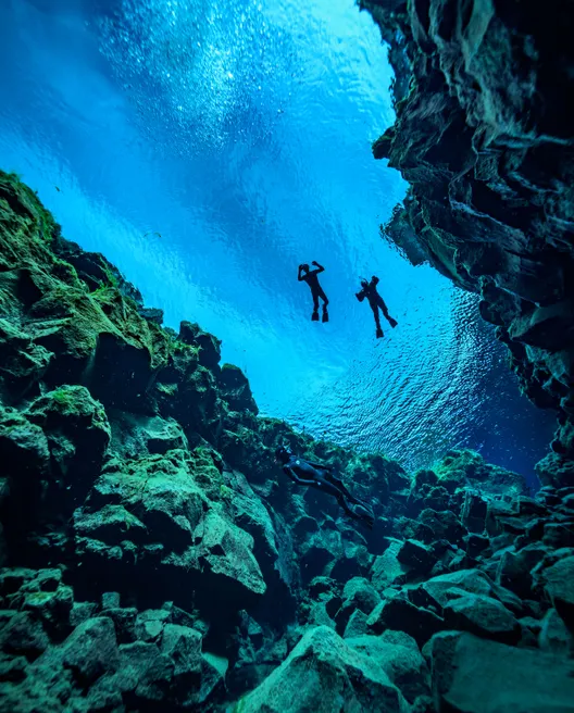 Sillouettes of two freedivers up on the surface of Silfra's vibrant blue waters between rocky fissure walls.