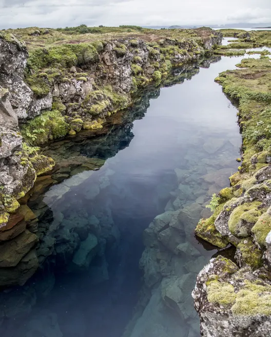 A long, narrow water- filled rift surrounded by moss covered rocks reflects sun on a partially cloudy day