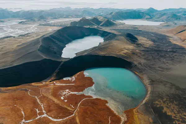 Vibrant rusty-orange terrain leads to a turquoise colored lake in a volcanic crater, in front of a second crater lake.