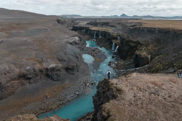 A person stands at a brown canyon edge overlooking the vibrant blue river flowing through it, with waterfalls pouring over the sides. 