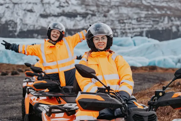 Two ATV riders in bright orange coveralls pose excitedly in front of a vibrant blue glacial outlet in winter.