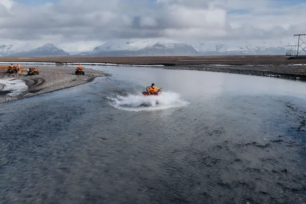 An ATV rider makes a big splash as they cross a wide, clear river, the sun gives it a light blue appearance.