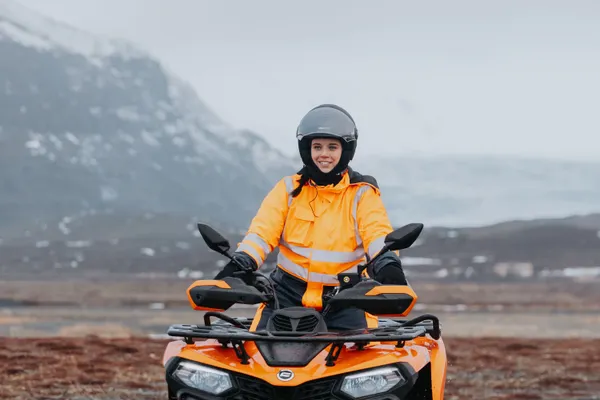 A woman on an ATV rides on a gravel track on a misty morning with mountains in the background.