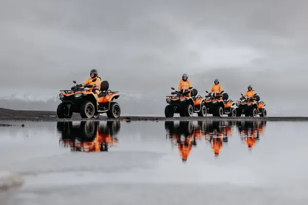 The reflections of four people on ATVs are seen in a small, still pond as they ride past on a cloudy day.