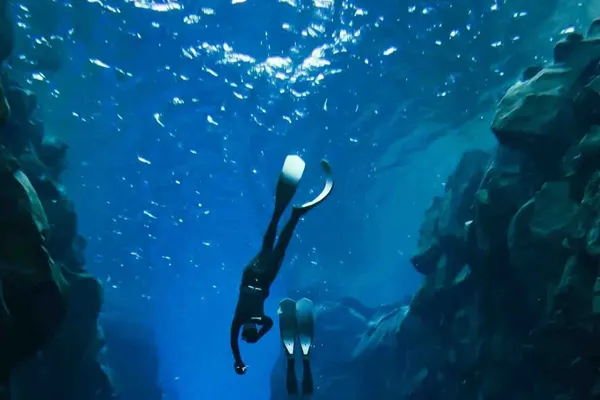 Two freedivers dive deeper into a fissure filled with clear, deep blue water.