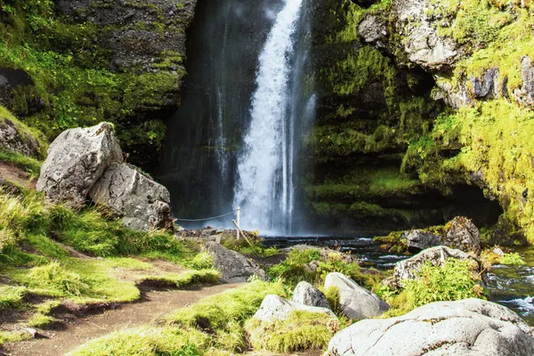 A partially concealed waterfall flows between layered rock formations covered in green moss on a sunny day.