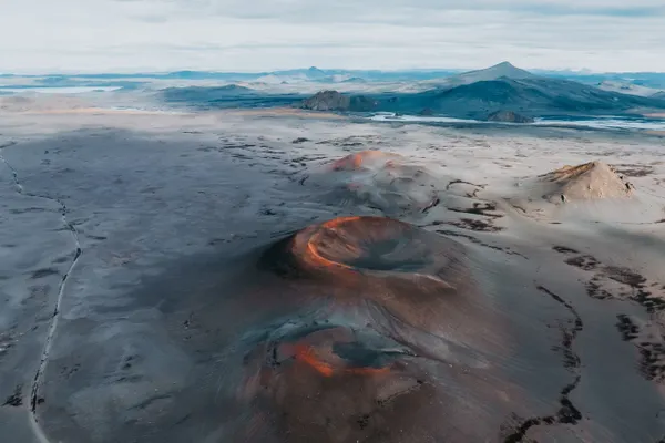 Three volcanoes with orange rims in the middle of ash- brown flat land, with mountains in the distant background.